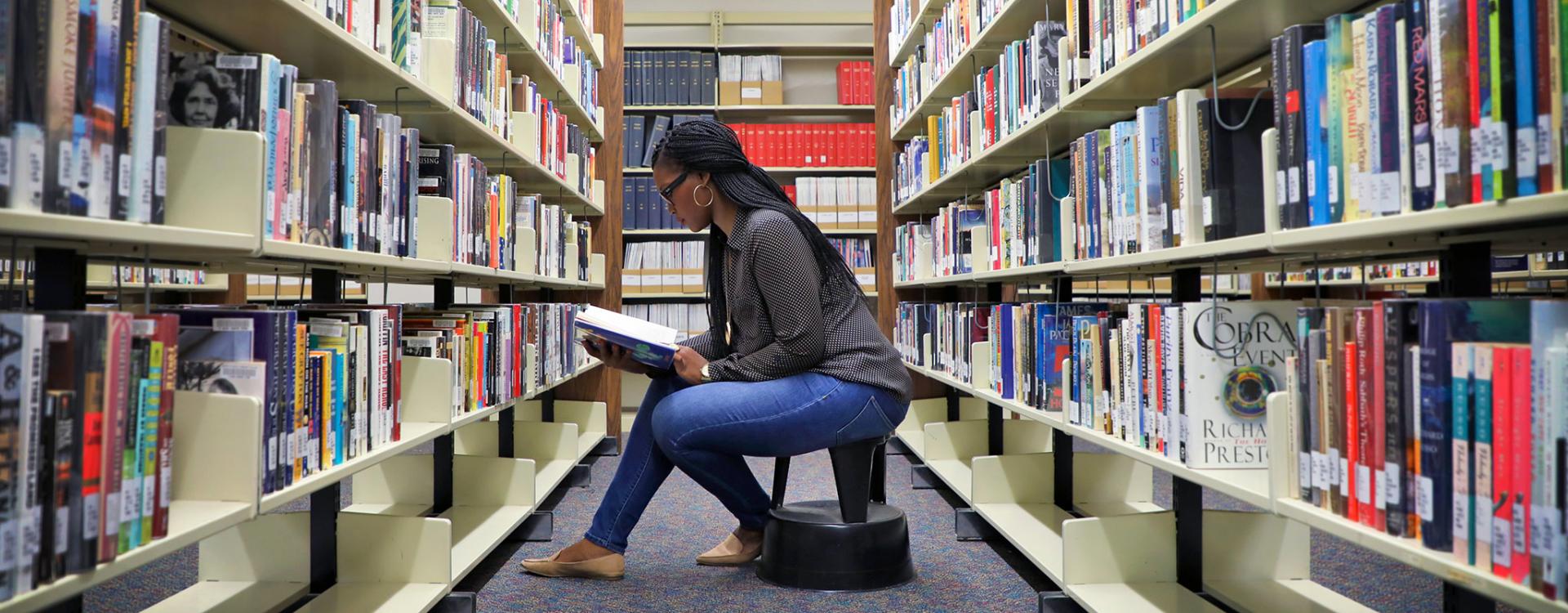 female student sitting on a step stool reading a book she just pulled from the library shelf